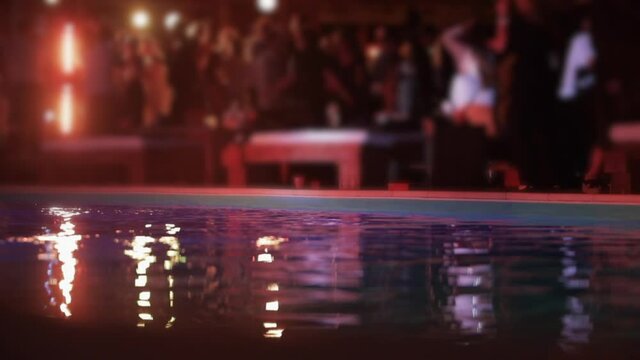 A pool with silhouettes of dancing people and concert lights reflected in the water, people dancing in the background of the pool out of focus.