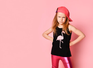 Unhappy little girl standing with hands on hips and bending brows looking at camera. Three quarter length portrait isolated on pink, copy space