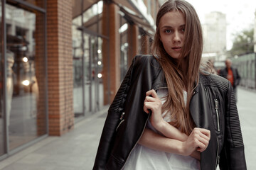 wide angle medium shot portrait young woman with long blonde hair wearing casual black leather jacket draped over her shoulders looking and posing at the camera