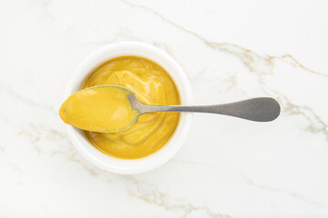 Mustard sauce in a bowl and spoon on white marble table. Top view