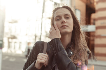 Portrait confident young woman on a blurred building background. Model wearing black eco-leather jacket and backpack