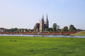 Fototapeta na wymiar Cuijk, Netherlands - August 9. 2020: View over green pasture and river maas on cityscape of dutch village with cathedral (focus on church)