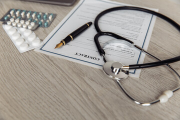 Modern doctor's office desk. Stethoscope with pills and healthcare contract on wooden table close-up.