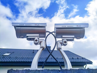 Two security cameras on the fence against the roof of the house and the blue sky with clouds.