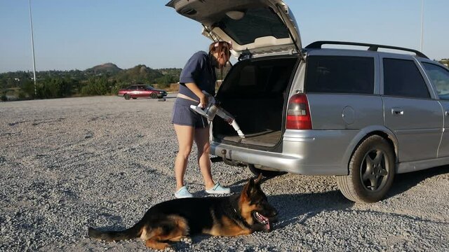 Young Woman Vacuuming Car Trunk Using A Vacuum Cleaner Removing Dirt And Dust From Car Trunk Outdoors At Sunset With Her Pet Dog