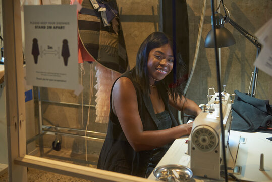 Young female designer working at sewing machine behind plexiglass glass in a cover-19 safe workplace.