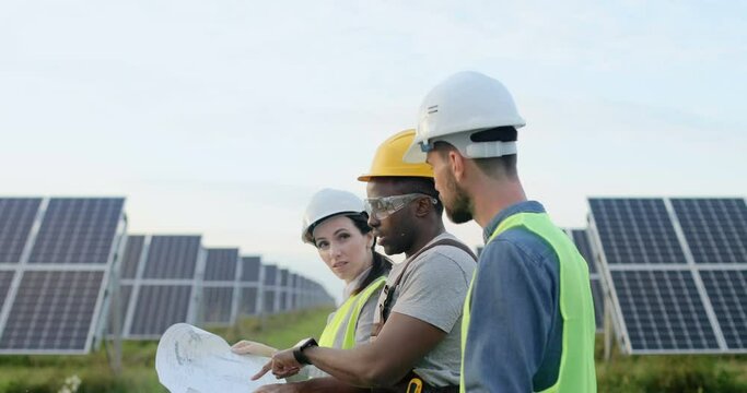 Three employees of alternative power plant walking and talking about scheme of solar panels. Team work. Group of specialists. Camera follows people.