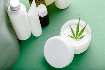 Hemp moisturizing cream in white jar with CBD oil cannabis leaf and set skin care cosmetics on green background