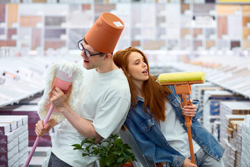 cheerful married caucasian couple have fun in the market, young caucasian man and woman hold mops...