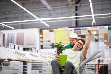 positive consultant drive customer in trolley in market aisle, they have fun and laugh, crazy customer with green plant in hands enjoy shopping