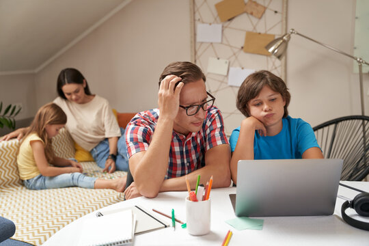 Worried father helping his little son with homework while mother and daughter sitting on sofa and playing with pazzles