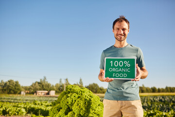 Portrait of smiling man holding ORGANIC FOOD sign and looking at camera while standing at vegetable plantation outdoors in sunlight, copy space