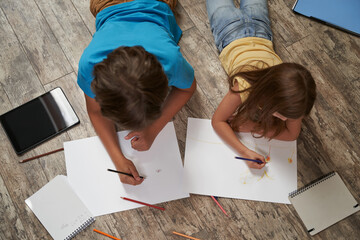 Siblings playing together at home. Top view of little boy and girl lying on the wooden floor and drawing on white sheets of paper with colorful pencils