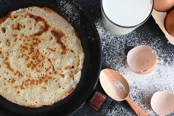 Hot pancake in black pan on black table with flour, milk and eggs