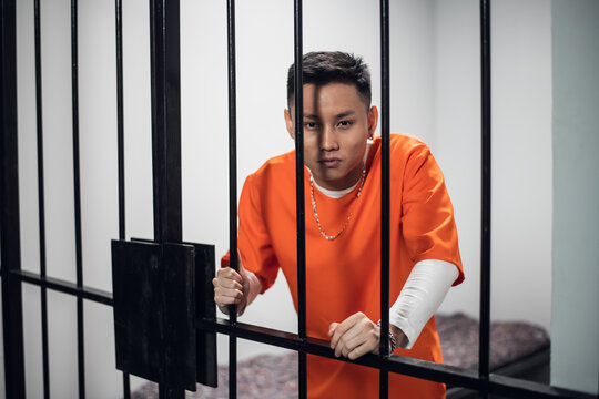 A young asian criminal poses for a photo against the bars in his cell.