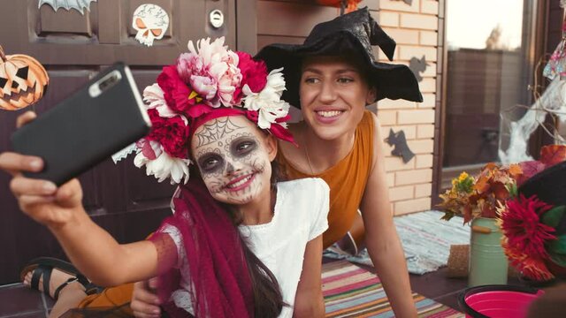 Handheld shot of happy young woman and kids in halloween costumes taking selfie on porch of house