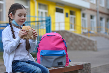 A schoolgirl with a protective medical mask, with a backpack, eating delicious pastries for lunch near the school. Back to school. New normal. - Powered by Adobe