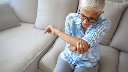 Fototapeta premium Senior woman with arm pain.Old female massaging painful hand indoors. Old woman hand holding her elbow suffering from elbow pain. Senior woman suffering from pain in hand at home. 