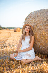 Attractive barefoot girl in white dress and a straw hat near the bale of straw in the field after the bread harvest
