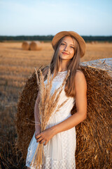 Young blond girl leaning on the bale of straw and holding a bouquet of dried flowers catching last warm sun rays of a setting sun