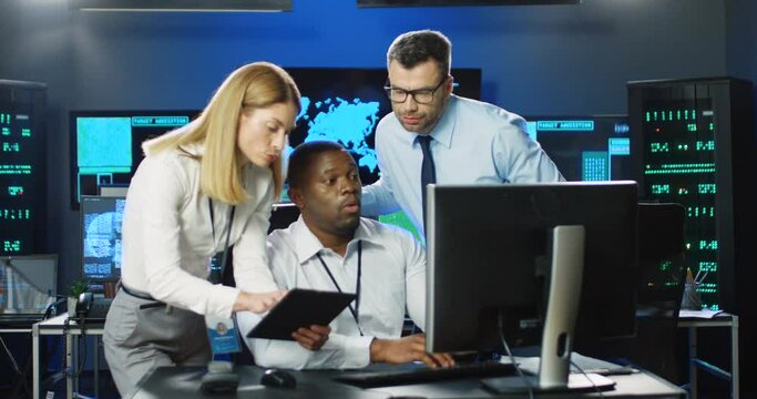 Caucasian woman with tablet asking for advice. African American boss man sitting at computer in database security office. Female males colleagues discussing info in cyber center Mixed-races Muliethnic