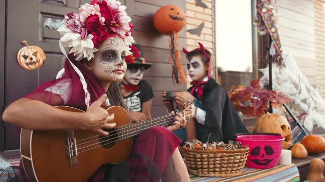 Handheld shot of cute girl wearing scary makeup and halloween costume sitting on steps on front porch and playing guitar. Boys in costumes sitting on background and chatting