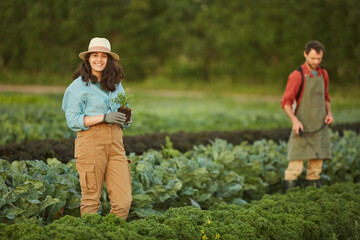 Portrait of two people working in field at vegetable plantation, focus on young woman smiling at camera in foreground, copy space