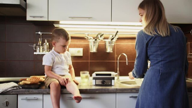Young mother and her son are making waffles in waffle maker at home kitchen. Woman washing dishes while waffles cooking