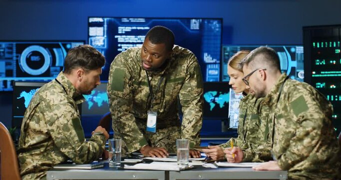 African American general heading at mixed-races militarian council at table in uniforms, deciding war or defense tactics in dark monitoring room. Multiethnic men and woman officers discussing warfare.
