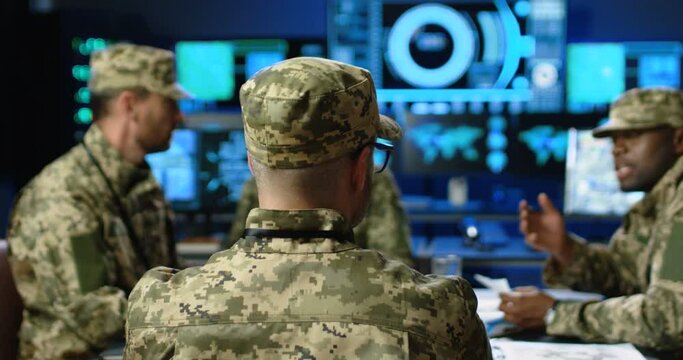 Rear of general with mixed-races militarian council sitting at table, deciding war or defense tactics in dark monitoring room. Back view of multiethnic people, officers talking, discussing terrorism.