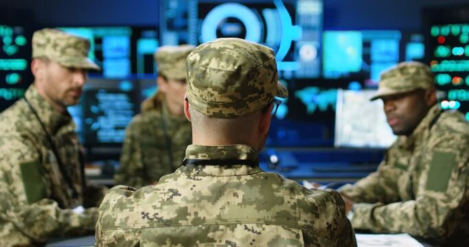 Rear of army chief with mixed-races militarian council sitting at table, deciding war or defense tactics in dark monitoring room. Back view multiethnic generals, officers talking, discussing terrorism