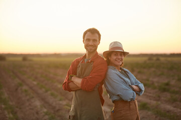 Waist up portrait of young farmers couple posing back to back while standing in field at sunset and smiling at camera, copy space