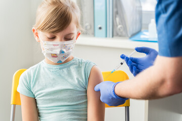 Little girl in face mask in doctor's office is vaccinated. Syringe with vaccine for covid-19...