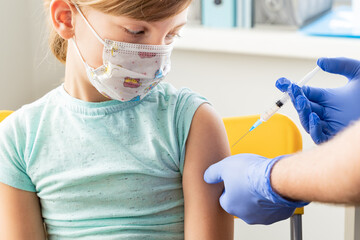 Little girl in face mask in doctor's office is vaccinated. Syringe with vaccine for covid-19...