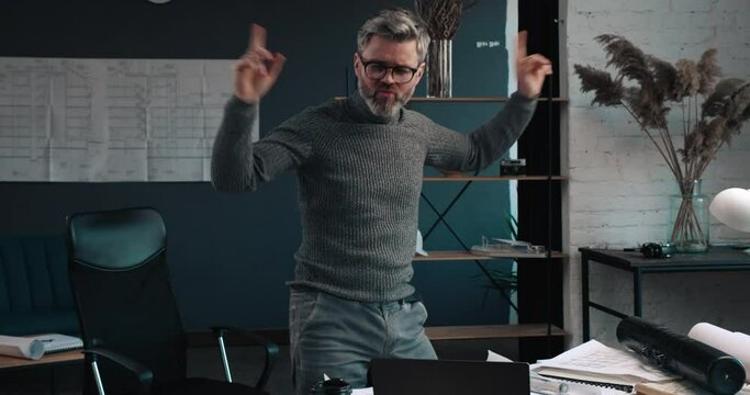 Handsome Architect making signs of Success and dancing because of successful deal. Looking satisfied and Happy with his job. Working at Modern office and making drawings on the laptop.