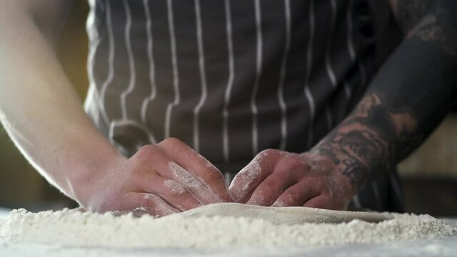 Close up of Male Tattoed Hands rolling out Dough for coking Pizza. Baker Preparing Dough for Pizza on a special Table before Baking. Traditional pizzeria Kitchen. Italian Cucine.