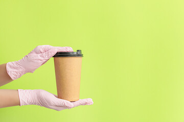 Hands in protective gloves and with cup of coffee on color background