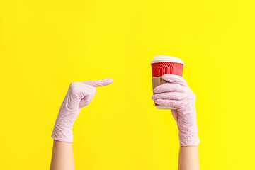 Hands in protective gloves and with cup of coffee on color background