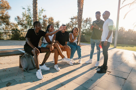 Group Of Young Modern African Black Friends Happily Sitting Together On The Bench Of The Stairs In Bright Sunlight Enjoying Summer Vacation