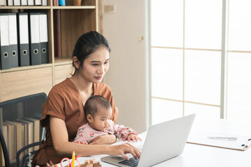 Happy mother working online with a laptop holding her baby daughter at home office. Smiling single...