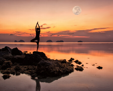 Silhouette of a young woman standing on the rocks by the sea in a yoga pose against the backdrop of a spectacular sunset and a huge rising full moon on a summer evening.