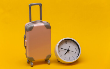 Time to travel. Mini plastic travel suitcase and clock on yellow background. Minimal style