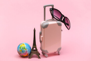 Traveled concept. Mini plastic travel suitcase with sunglasses and statuette of the Eiffel Tower, globe on pink background.