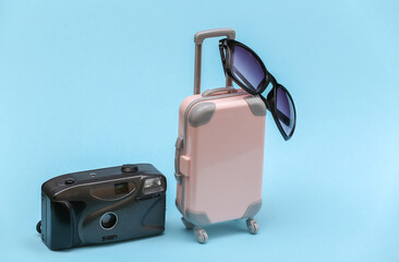 Travel concept. Mini pink plastic travel suitcase with camera, sunglasses on blue background. Minimal style