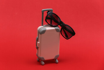 Travel or trip concept. Mini plastic travel suitcase with sunglasses on red background. Minimal style