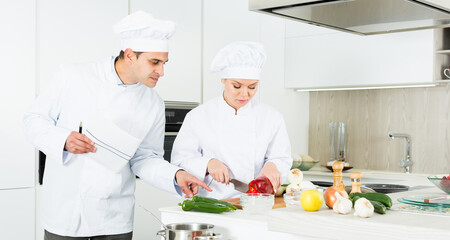 Positive female and male young cooks with paper recipe in uniform working on kitchen