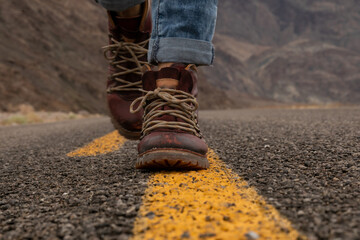 woman in jeans and brown boots walking on a yellow line on the empty road, closeup to feet