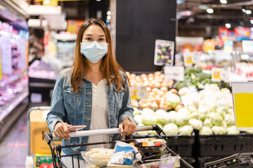 Young Asian women wearing surgical mask buying fresh vegetable in a grocery store at the supermarket during coronavirus outbreak, New normal lifestyle concept