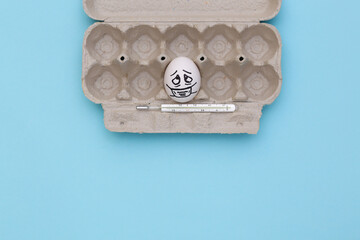 One egg face in medical mask with thermometer in tray on blue background. Covid-19 pandemic. Top view