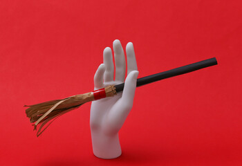 White mannequin hand holding mini witch's broom on red background. Halloween concept
